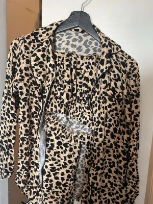 Leopardmönstrad pyjamas i två delar - Snygg pyjamas i två delar med leopardmönster. Setet består av en långärmad skjorta med krage och svarta kantdetaljer samt matchande byxor med resår i midjan. Perfekt för dig som vill ha en trendig och bekväm look hemma.