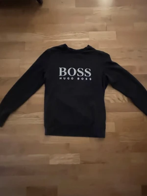 Svart tröja från Hugo Boss - Snygg svart långärmad tröja från Hugo Boss med stort vitt BOSS-tryck och HUGO BOSS-text på bröstet. Klassisk rund halsringning och enkel design som passar till det mesta. Perfekt för dig som gillar stilrena plagg med tydlig logga.