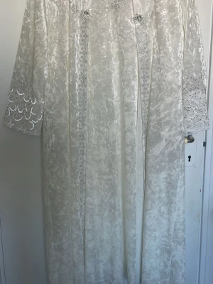 Vit långklänning med spetsdetaljer - Elegant vit långklänning med vackra spetsdetaljer och glittriga inslag på ärmar och längs framsidan. Klänningen har lång ärm och är tillverkad i ett mjukt, glansigt material som ger en lyxig känsla.