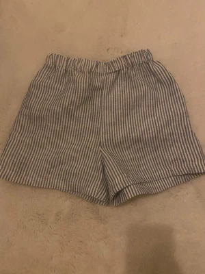 Randiga shorts i linne-look - Säljer ett par avslappnade shorts med blå och vita ränder. De har elastisk midja och är tillverkade i ett material som liknar linne, perfekt för varma dagar. Passformen är loose och de har en klassisk, enkel design utan extra detaljer. Passar de som har strl xxs/xs/s
