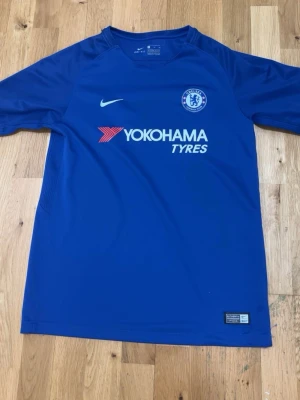 Chelsea FC matchtröja blå Nike - Chelsea FC matchtröja i blått från Nike med klubbmärke och sponsortryck 'Yokohama Tyres' på bröstet. Tillverkad i lätt och ventilerande material, perfekt för fotbollsträning eller match. Klassisk rund hals och korta ärmar.