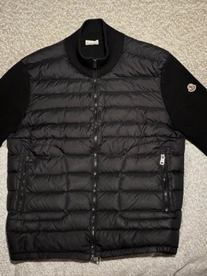 Svart dunväst/ cardigan från Moncler - Stilren svart dunväst från Moncler med quiltad design och dragkedja framtill. Västen har två praktiska dragkedjefickor och en ribbad halskrage. För dig som är osäker på storlek så passar den dig som är mellan 170 ~185cm.       Passformer är perfekt och den fungerar till T-shirts eller hoodie under vilket gör att man kan använda den till alla tillfällen. Priset kommer att ligga på 2000kr till en början men det kan diskuteras.
