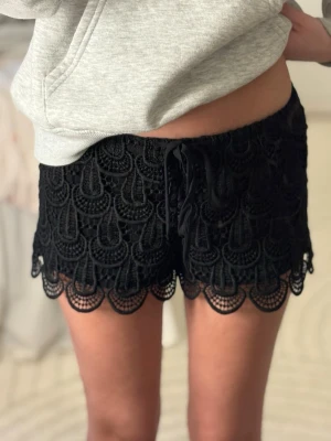 Svarta spetsshorts med snörning - Snygga svarta shorts med virkad spets och dekorativ snörning i midjan. Shortsen har ett unikt mönster och vågig kant nertill som ger en extra detalj. Perfekta till sommaren för en trendig och avslappnad look. 