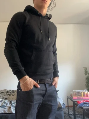 Svart hoodie med ficka framtill - En stilren svart hoodie med huva och dragsnören. Hoodien har en stor känguruficka framtill och ribbade muddar vid ärmslut och nederkant. Perfekt för en avslappnad och cool look. Materialet känns mjukt och bekvämt mot huden.