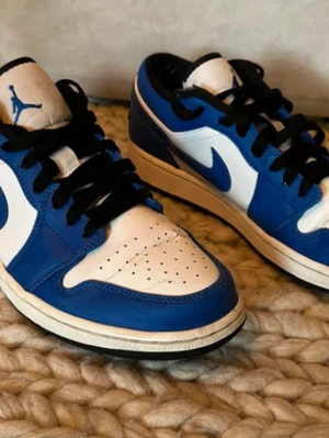 Nike Air Jordan 1 Low blå/vit - Snygga Nike Air Jordan 1 Low sneakers i blått och vitt med svarta skosnören och klassisk swoosh på sidan. Skorna har en låg silhuett och är tillverkade i läder med perforerad tåbox för extra ventilation. Perfekta för dig som gillar streetwear och sportig stil.