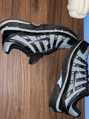 Nike P-6000 svart och grå sneakers - Nike P-6000 sneakers i svart och grått med vit sula. Skorna har en sportig design med mesh och syntetmaterial, tydlig snörning och den klassiska Nike Swoosh-loggan på sidan. Perfekta för en streetwear-look.