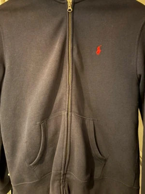 Mörkblå hoodie från Ralph Lauren - Mörkblå hoodie med dragkedja från Ralph Lauren.  Perfekt för en avslappnad och stilren look. Mjuk bomullsblandning som känns skön mot huden.