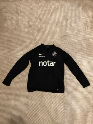 Svart AIK träningströja Nike - Svart långärmad träningströja från Nike med AIK-logga på bröstet och tryckta sponsorer. Tröjan har halv dragkedja framtill och grå detaljer längs ärmarna. Perfekt för fotbollsträning eller gym. Materialet är troligtvis polyester för bra andningsförmåga.