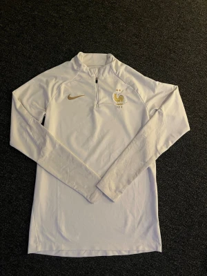 Vit Nike Frankrike träningströja - Vit långärmad träningströja från Nike med Frankrikes landslagslogga och guldigt Nike-märke på bröstet. Tröjan har halv dragkedja framtill och är tillverkad i ett lätt och stretchigt material, perfekt för sport och träning.