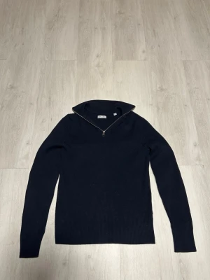 Mörkblå stickad tröja med dragkedja - Snygg mörkblå stickad tröja.  Med ribbad struktur och dragkedja vid halsen. Tröjan har lång ärm och en hög krage som kan vikas ner. Perfekt för dig som gillar stilrena och klassiska plagg. Storlek M. 