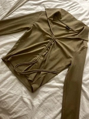 Beige långärmad topp med snörning - Trendig beige topp med långa ärmar och snygg snörningsdetalj framtill. Toppen har en djup v-ringning och krage som ger en cool look. Perfekt att styla med jeans eller kjol för en chic outfit.