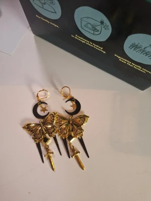 Guldiga statement örhängen med fjärilar - Unika örhängen i guld- och svartfärgad metall med stora detaljerade fjärilar, svarta månar med små stjärnor, samt hängande svärd och spjut. Perfekt för dig som gillar iögonfallande accessoarer med mystisk och alternativ vibe.
