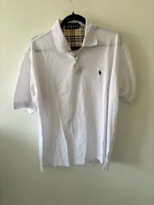 Vit pikétröja från Ralph Lauren. Passar även L - Klassisk vit pikétröja från Ralph Lauren med korta ärmar och krage. Tröjan har knappslå med rutigt tyg på insidan och den ikoniska loggan broderad på bröstet. Perfekt till jeans eller shorts för en clean look.