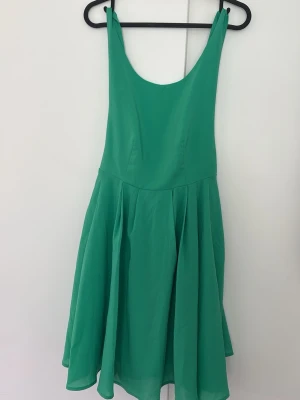 Grön ärmlös klänning med knyt - Snygg grön klänning med vidd i kjolen och djup urringning både fram och bak. Klänningen är ärmlös och har ett dekorativt knyt i ryggen. Perfekt för sommarens alla tillfällen och tillverkad i ett lätt, mjukt tyg.