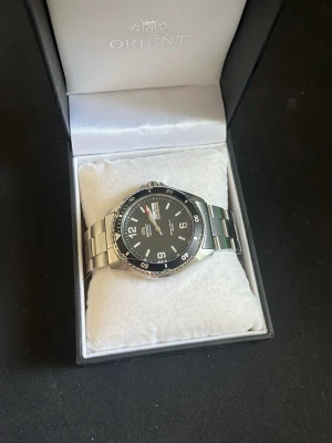 Orient svart/silver herrklocka - Stilren klocka från Orient med svart urtavla, tydliga vita siffror och datumvisning. Boetten och länken är i silverfärgat metall, vilket ger en klassisk look. Perfekt för dig som gillar en sportig men ändå elegant stil. Den är nerkortat med tre länkar, men kommer med fler om man vill lägga till. Klockan är varmsamt använd och är som ny. Fråga på vid minsta fundering!