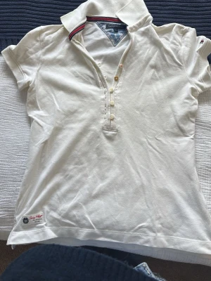 Vit polotopp från Tommy Hilfiger - Vit polotopp från Tommy Hilfiger med klassisk krage och knappar framtill. Toppens krage har en snygg detalj med röd och marinblå rand. Tillverkad i mjuk bomull och har korta ärmar. Perfekt för en elegant  look, passar jätte bra med ett par lågmidjade jeans