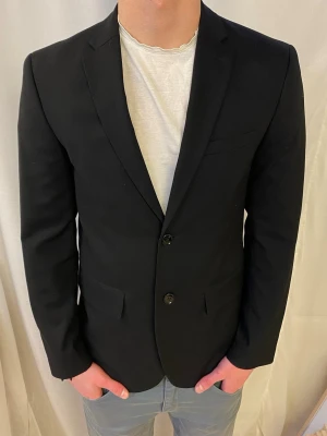 Svart slim fit kavaj från H&M - Stilren svart kavaj från H&M i slim fit-modell. Kavajen har två knappar framtill, klassiska slag och fickor med lock. Perfekt för dig som vill ha en enkel och snygg look. Materialet är mjukt och följsamt för en bekväm passform.