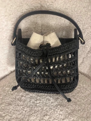 Svart flätad handväska med axelrem - Snygg svart handväska i flätad design med både handtag och justerbar axelrem. Väskan har en innerpåse i beige tyg med dragsko och detaljer i guld. Perfekt för dig som gillar trendiga accessoarer och vill ha något unikt till din outfit.