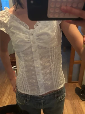 Vit broderad topp med knappar - Supersöt vit topp med broderade hål, volangärmar och dekorativa knappar framtill. Figurnära passform och lätt rynk vid bysten. Perfekt till jeans eller kjol för en fräsch look. Materialet känns luftigt och somrigt.