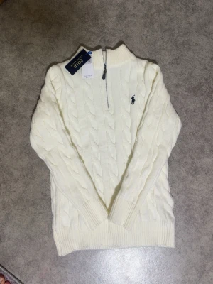 Vit kabelstickad tröja Polo Ralph Lauren - Vit kabelstickad tröja från Polo Ralph Lauren med halv dragkedja och broderad logga på bröstet. Tröjan har ribbade muddar och hög krage, perfekt för en stilren look. Tillverkad i mjukt material för extra komfort.