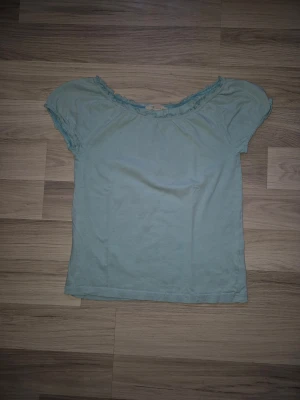 Ljusblå topp med volangkant - Söt ljusblå topp med korta ärmar och volangdetalj längs den vida, rundade halsringningen. Perfekt för dig som gillar en enkel men ändå feminin stil. Tillverkad i mjukt bomullsmaterial som känns skönt mot huden.