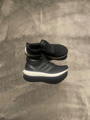 Adidas Ultra Boost svarta sneakers - Svarta Adidas Ultra Boost sneakers med stickad ovandel och vita Boost-sulor. Skorna har klassisk tre-randig design på sidorna och svart hälkappa. Perfekta för dig som gillar sportig och stilren look.