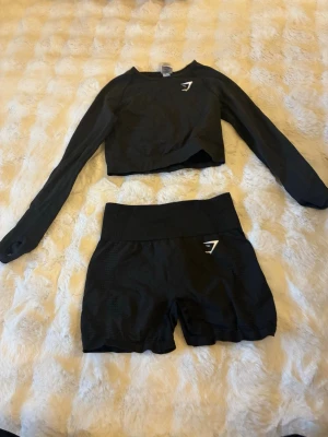 Svart träningsset från Gymshark - Säljer ett svart träningsset från Gymshark med långärmad crop top och matchande shorts. Båda plaggen har Gymshark-loggan i vitt och är tillverkade i ett stretchigt, bekvämt material som passar perfekt för gymmet eller löpning.