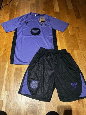FC Barcelona summer set purple/black - Säljer ett snyggt träningsset från FC Barcelona med lila t-shirt och svarta shorts. Tröjan har svarta detaljer på sidorna, klubbmärke och Spotify-logga framtill. Shortsen är svarta med lila detaljer och klubbmärke. Perfekt för fotbollsträning eller gymmet.