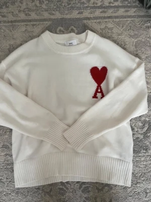 Ami Paris, strl s, superbra skick  - Vit stickad tröja från Ami Paris med röd hjärtlogga och bokstaven A på bröstet. Tröjan har rund halsringning, ribbade muddar och nederkant. Perfekt för dig som gillar stilrena plagg med en twist.