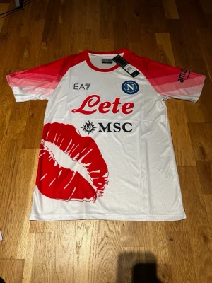 Napoli valentines day special edition kit - Cool Napoli fotbollströja från EA7 i vitt med röda detaljer och stort rött kyssmärke framtill. Tröjan har röda och rosa ärmar, rund hals och tryckta loggor från Lete och MSC. Perfekt för dig som vill sticka ut på match eller träning.