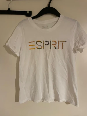 Vit t-shirt från Esprit med tryck - Vit t-shirt från Esprit med färgglatt logotyptryck på bröstet. Klassisk rund halsringning och korta ärmar. Tillverkad i mjukt bomullsmaterial som känns skönt mot huden. Perfekt för en avslappnad och stilren look.