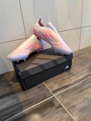 Adidas X Speedflow rosa fotbollsskor - Snygga Adidas X Speedflow fotbollsskor i vitt och rosa med svarta detaljer. Skorna har en strömlinjeformad design och är tillverkade i lätt syntetmaterial för maximal fart på planen. Perfekta för dig som vill sticka ut och spela snabbt.