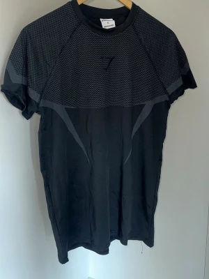 Svart träningströja från Gymshark - Snygg svart träningströja från Gymshark med diskreta grå detaljer och meshmönster upptill. T-shirten har korta ärmar och är tillverkad i ett lätt och stretchigt funktionsmaterial som andas, perfekt för gymmet eller löprundan. OBS rep passar S är en muselfit, kan även passa M lite tajtare då! 