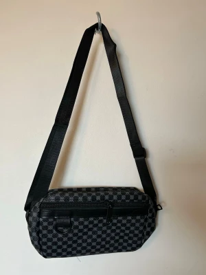 Svart axelväska med rutigt mönster - Snygg svart axelväska med justerbar rem och rutigt mönster i grått och svart. Väskan har en dragkedja framtill och en ringdetalj för extra stilpoäng. Perfekt storlek för det viktigaste och enkel att matcha till olika outfits.