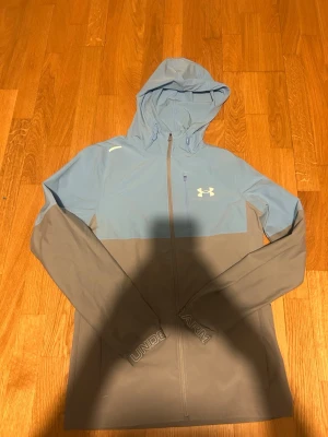 Blågrå vindjacka från Under Armour - Snygg tvåfärgad vindjacka från Under Armour i ljusblått och grått. Jackan har huva, hel dragkedja framtill, bröstficka med dragkedja och logga på bröstet. Ärmslut med broderad text och lätt material som passar perfekt för blåsiga dagar.