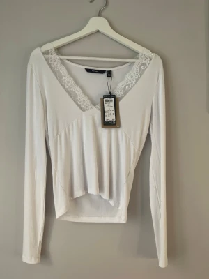 Vit spetsdetaljerad topp Vero Moda - Vit långärmad topp från Vero Moda med v-ringning och spetsdetaljer runt halsen. Tillverkad i mjuk bomullsmix och har en lätt croppad passform. Perfekt för dig som gillar en feminin och stilren look.