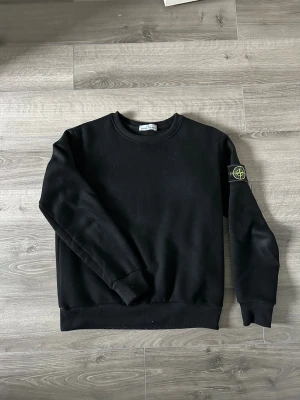 Svart stone Island tröja  - Svart crewneck sweatshirt från Stone Island med klassisk rund hals och ribbade muddar. Ikonisk logopatch på vänster ärm ger en streetwear-vibe. Perfekt till jeans eller cargo för en clean och stilren look.