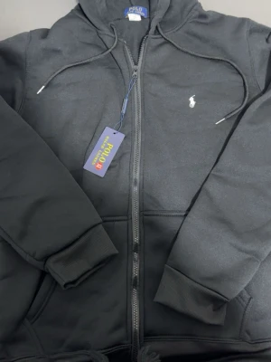 Svart hoodie från Polo Ralph Lauren - Svart hoodie med dragkedja från Polo Ralph Lauren. Klassisk design med vit broderad logga på bröstet, huva med dragsko och ribbade muddar. Tillverkad i mjukt bomullsmaterial för en skön och avslappnad look. Byxor följer med också helt ny dress