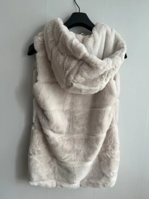 Vit fuskpälsväst med luva - Beige/vit fuskpälsväst från Amisu med stor mysig luva. Västen är gjord i mjukt, fluffigt material. Använd 1 gång, ser helt ny ut. 