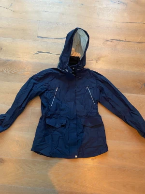 Mörkblå vårjacka från Didriksons - Snygg mörkblå vårjacka från Didriksons med huva och dragkedja. Jackan har två bröstfickor med dragkedja och två stora fickor framtill med lock. 100% vattentät och perfekt för regniga dagar. Klassisk design med justerbar midja och detaljer i silver.  Inköpt för 1800kr väldigt gott skick 