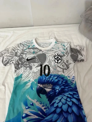 Vit och blå Brasilien fotbollströja - Säljer en unik fotbollströja från Nike med Brasilien-tema. Tröjan är vit med ett stort tryck av en blå papegoja och grafiska detaljer i grått och turkost. Nummer 10 på bröstet och CBF-logga. Perfekt för dig som vill sticka ut på planen!
