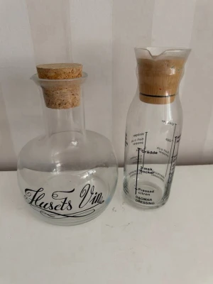 Två glasflaskor med kork och text - Säljer två stilrena glasflaskor. Den ena har texten 'Husets Vin' och en korkpropp, perfekt som vinkaraff. Den andra har måttangivelser och trälock, smidig för dressing eller sås. Båda är genomskinliga och har snygg design som passar i köket.
