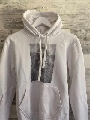 The Cooler Elephant Hoodie - Säljer en vit hoodie med stor magficka och huva med dragsko. På bröstet finns ett rektangulärt grafiskt tryck i svartvitt. Hoodien har ribbade muddar vid ärmslut och nederkant. Perfekt för dig som gillar minimalistisk streetwear.
