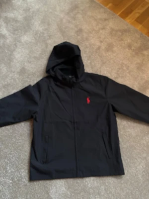 Svart vindjacka från Polo Ralph Lauren - Snygg svart vindjacka från Polo Ralph Lauren med huva och dragkedja framtill. Jackan har det klassiska röda Polo-logotypen broderad på bröstet och två sidofickor. Tillverkad i lätt och tåligt material, perfekt för blåsiga dagar.