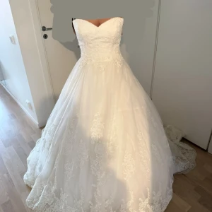 Vit prinsessklänning med spetsdetaljer - Drömmig vit brudklänning i prinsessmodell med vacker spets upptill och längs kjolen. Klänningen har hjärtformad urringning och en lång, vid kjol som ger en elegant siluett. Perfekt för dig som vill känna dig som en prinsessa på din stora dag.