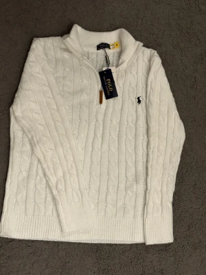 Ralph Lauren half zip - Kabelstickad vit tröja från Polo Ralph Lauren med halv dragkedja och ribbade muddar. Klassisk svart logga broderad på bröstet och hög krage. Perfekt för dig som gillar stilrena och tidlösa plagg i mjukt material.
