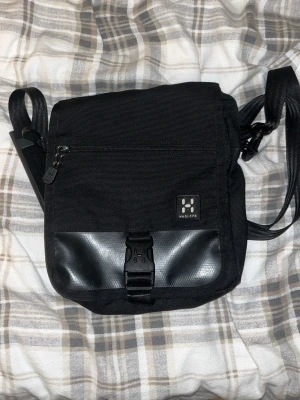 Svart axelväska från Haglöfs - Snygg svart axelväska från Haglöfs med justerbar rem, flera fack och smart spänne framtill. Väskan har en kombination av slitstarkt tyg och detaljer i konstläder. Perfekt för dig som vill ha en praktisk och stilren väska med många förvaringsmöjligheter. Väskan har något typ av klister inne i facken men det försvinner lätt. Väskan är perfekt för sig som gillar väskor som är enkla och bära men som ändå bär stil. Priset är inte hugget i sten.