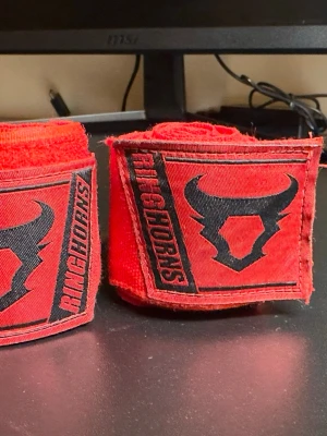 Röda boxningslindor från Ringhorns - Säljer ett par röda boxningslindor från Ringhorns med svart logga och text. Perfekta för träning inom boxning eller kampsport. Tillverkade i elastiskt material för bra stöd och komfort. Snygg design med tydlig logotyp på kardborrebandet.