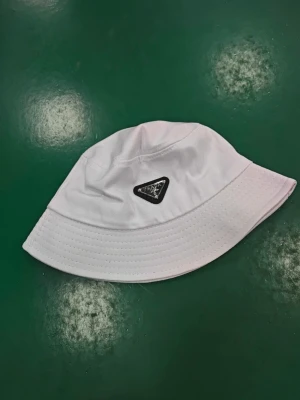 Vit bucket hat från Prada - HELT NY!!!! Säljer en stilren vit bucket hat från Prada med den klassiska svarta trekantsloggan framtill. Hatten har en mjuk, rundad form och är tillverkad i ett slätt tyg som ger en clean look. Perfekt accessoar för att lyfta din outfit.