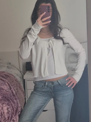 Vit ribbad topp/cardigan från Primark - Trendig vit ribbad topp/cardigan från Primark med lång ärm och croppad passform. Toppen har en öppen front med knytband och rundad urringning. Storlek 34-36. Perfekt att styla med jeans eller kjol för en snygg look. Skriv om prisförslag eller bilder ❤️🫶